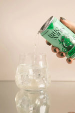 Little Saints can pouring a sparkling beverage into a glass filled with ice on a reflective surface.