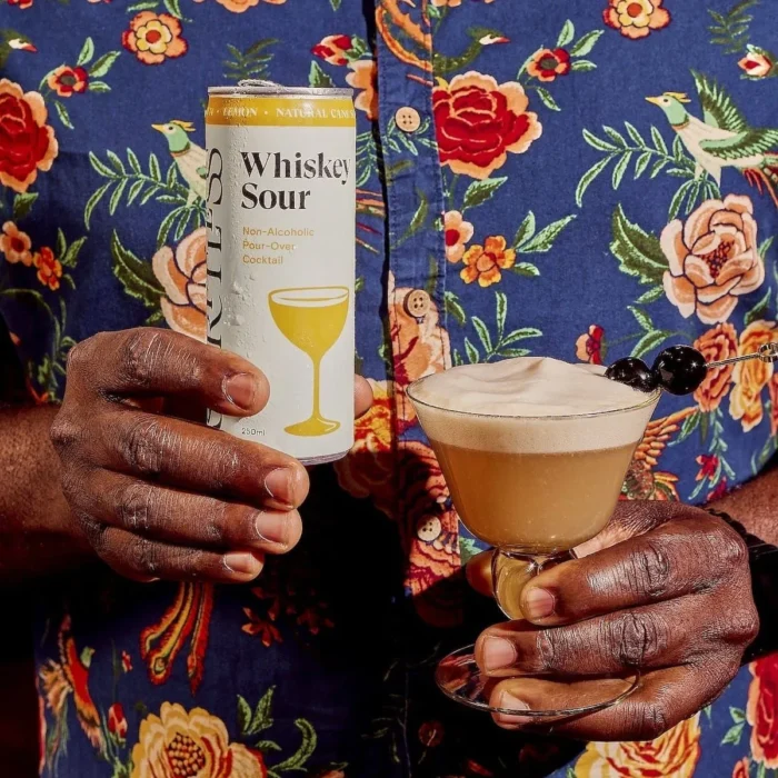 Person holding a Spiritless Whiskey Sour can with a garnished whiskey sour cocktail.