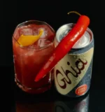 Ghia can with a chili pepper and a spicy red cocktail garnished with citrus peel on a dark background.