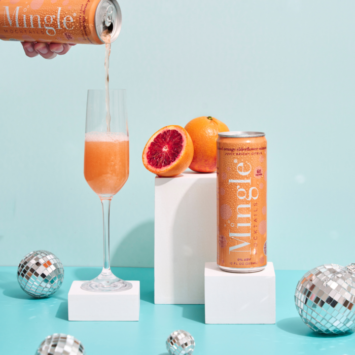 Pouring Mingle Mocktails Blood Orange into a flute glass, surrounded by fresh oranges and disco balls.