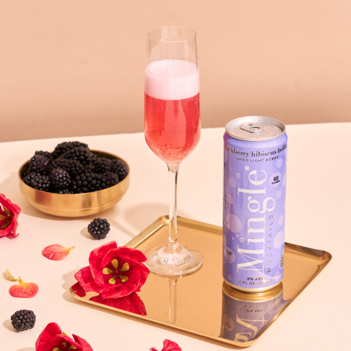 Flute glass filled with Mingle Mocktails Blackberry Hibiscus alongside the can, blackberries, and floral decor.