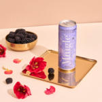 Mingle Mocktails Blackberry Hibiscus can on a gold tray with fresh blackberries and vibrant flowers.