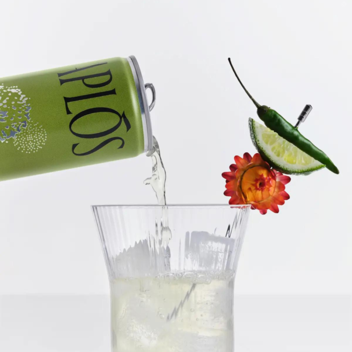 APLOS can pouring drink into a glass with lime, chili pepper, and flower garnish floating nearby.