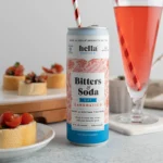 Hella Bitters Soda can with a striped straw next to a glass of red soda and tomato-topped appetizers.