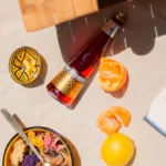 Non-alcoholic cocktail bottle with peeled citrus fruits and vibrant bowls of food on a sunlit table.