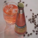 Sparkling non-alcoholic cocktail bottle with a vibrant iced drink and scattered coffee beans on a table.