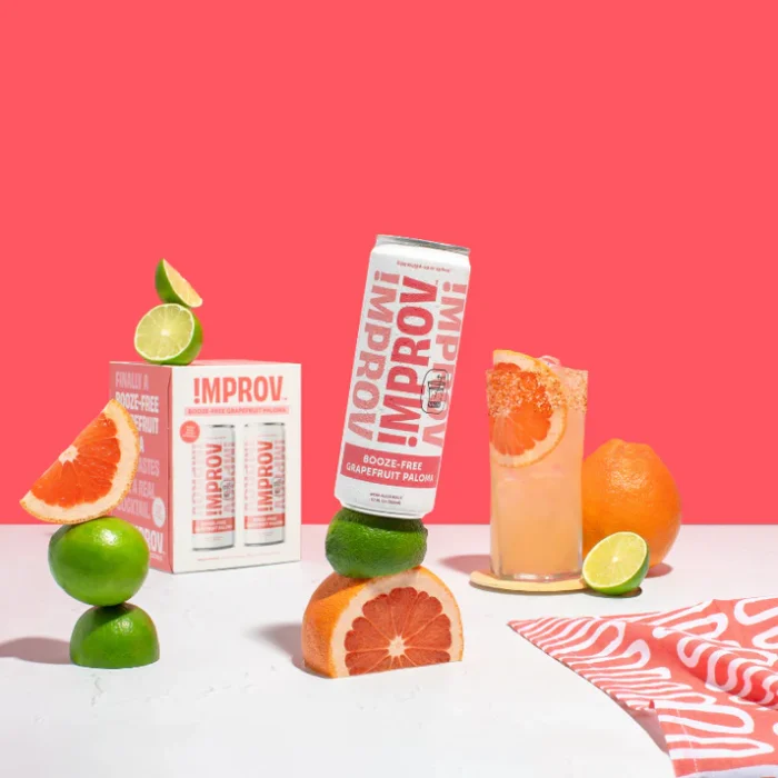 Improv citrus drink can with grapefruit, lime, and fresh cocktail setup against a vibrant pink background.