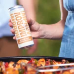 Person holding Improv ginger drink can over a sizzling pan of colorful grilled vegetables.