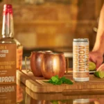 Improv ginger drink can with copper mugs, fresh lime, and mint on a wooden cutting board in a rustic setting.