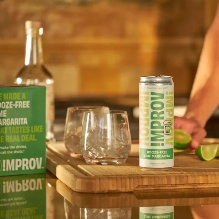 Improv Margarita drink can with fresh lime, glassware, and bottle on a wooden cutting board in a rustic kitchen.